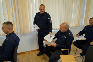 policjanci siedzą na krzesłach, słuchają organizatorów zawodów i przystępują do pisania testu